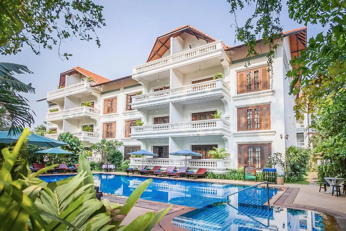 A three-story boutique hotel with white exterior walls, wooden window shutters, and spacious balconies overlooks a sparkling outdoor swimming pool surrounded by sun loungers, umbrellas, and lush tropical plants.