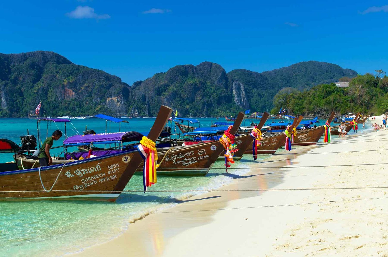 Colourful long-tail boats resting on a sandy southeast asian beach with turquoise water and a clear sky in the background